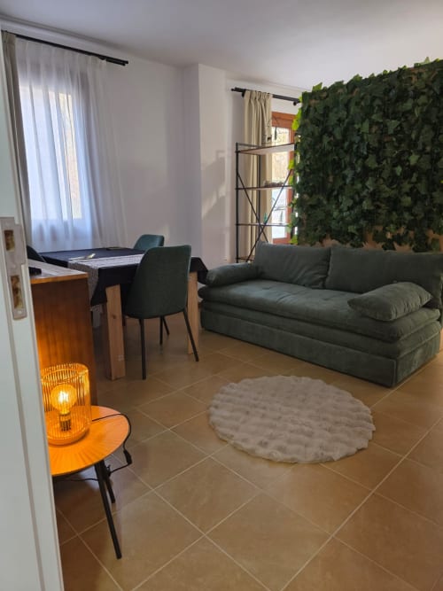 Moderno apartamento estudio en Apartamentos el Manantial de Chóvar con sofá gris, cama, cocina integrada y frondosa vegetación