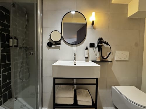 Salle de bains moderne avec lavabo blanc, meuble-vasque noir, miroir ovale et douche carrelée noire à l'Hôtel des Quatre Saisons