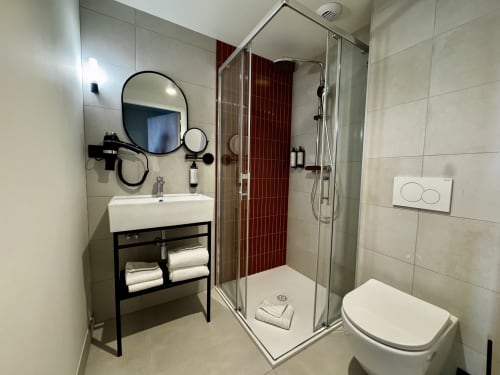 Salle de bains moderne de l'Hôtel des Quatre Saisons Guérande avec vasque noire, miroir rond, douche vitrée et carrelage rouge