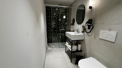 Salle de bain moderne de l'Hôtel des Quatre Saisons à Guérande avec douche carrelée noire, lavabo blanc, miroir ovale et toilettes blanches
