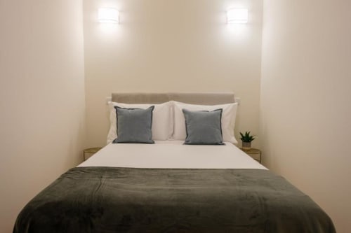 Quarto moderno no AgapeStay Porto com cabeceira bege, lençol branco, almofadas cinzentas, manta cinzenta escura e iluminação embutida