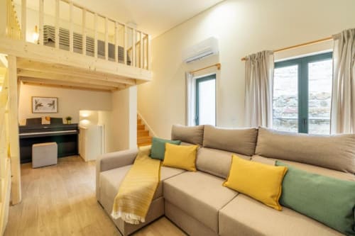 Sala luminosa com sofá bege, almofadas amarelas e verdes, quarto tipo loft, pisos de madeira e vista para o jardim na AgapeStay