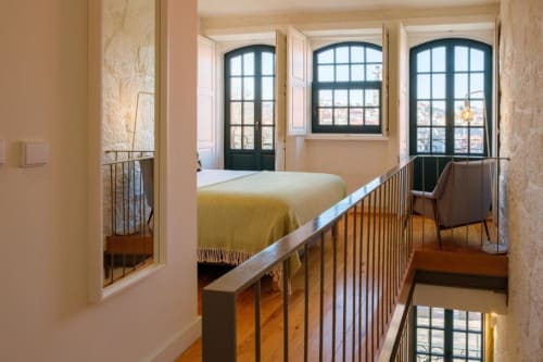 Quarto de apartamento duplex no Porto com mezanino, colcha amarela, assoalho em madeira e janelas em arco com vista para a cidade