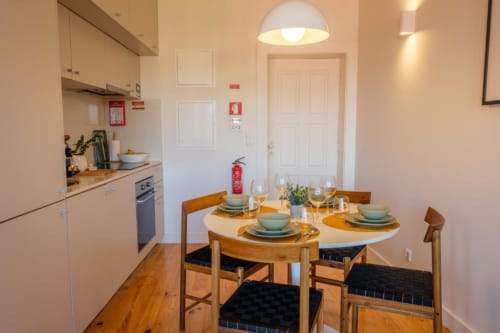Sala de jantar moderna em apartamento no Porto com mesa de madeira, utensílios de mesa e cozinha equipada com armários brancos.