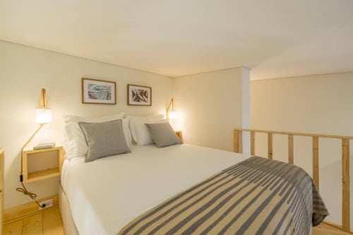 Quarto luminoso na AgapeStay Porto com cama queen, cabeceira em madeira, roupa de cama cinza listrada e iluminação aconchegante