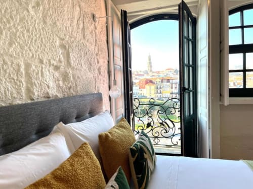 Quarto moderno com cabeceira cinza, almofadas douradas e portas de sacada pretas com vista para a colorida paisagem urbana do Porto e sua igreja