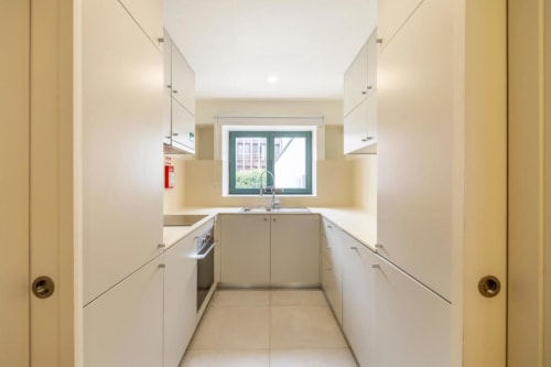 Cozinha galera moderna em apartamento no Porto com armários brancos, janela com vista para jardim, eletrodomésticos em aço inoxidável, e