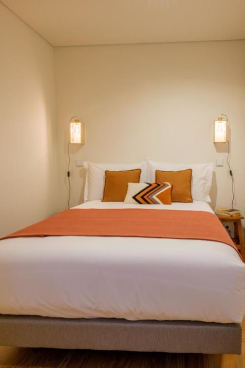 Quarto moderno no AgapeStay Porto com acentos em laranja, roupa de cama branca, iluminação em latão fixada à parede e decoração minimalista