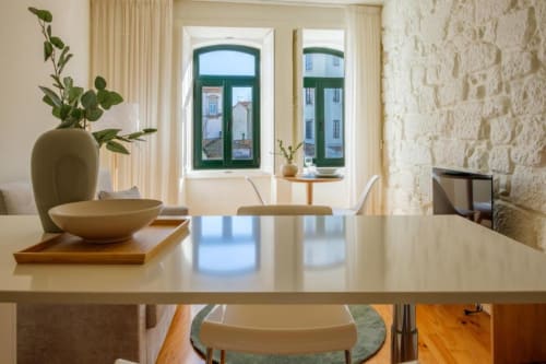 Sala de jantar iluminada em apartamento no Porto com janelas de moldura verde, parede de pedra, mesa de madeira e vista para o jardim