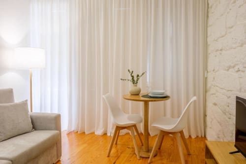 Elegante sala de estar em apartamento moderno com cadeiras brancas, mesa redonda em madeira, cortinas creme e piso de madeira no AgapeStay Porto
