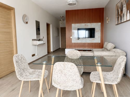 Modern dining area in Dakar apartment with glass table, upholstered chairs, and living room with TV mounted on rust-colored