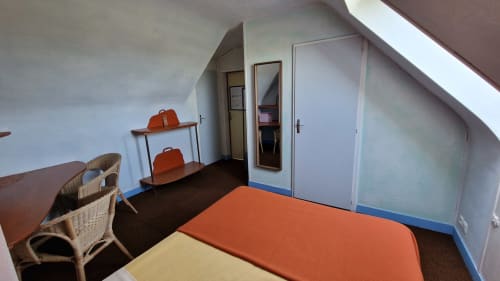 Chambre mansardée douillette à l'Hôtel Rose avec literie orange, fauteuil en rotin et plafond incliné à Beauvoir