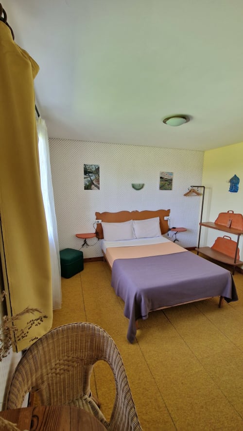 Chambre avec vue sur la rivière à l'Hôtel Rose à Beauvoir : lit double, mobilier en bois, rideaux jaunes et fauteuil en osier sur liège