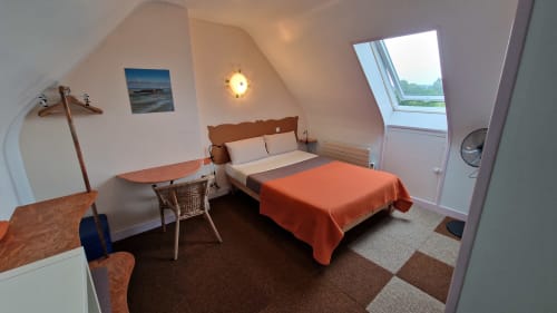 Chambre mansardée à l'Hôtel Rose avec couverture orange, vue rivière par lucarne, mobilier en bois et lumière chaleureuse à Beauvoir.