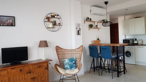 Moderno salón de apartamento en Nerja con silla de mimbre, mesa de comedor de madera, taburetes azules, cocina y TV de pantalla plana encendido