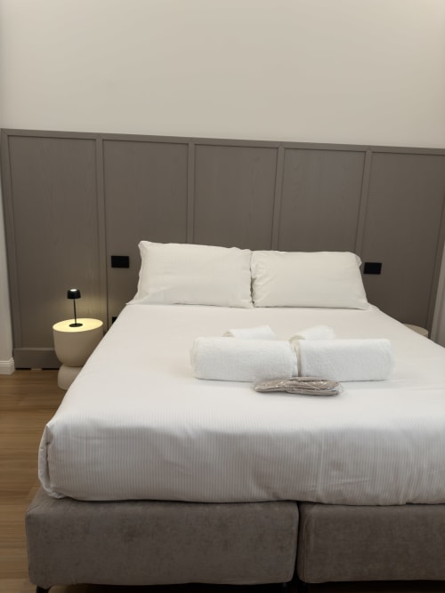 Camera elegante di Maison Riviera con biancheria bianca, testata grigia in tessuto, lampada comodino e asciugamani arrotolati sul letto