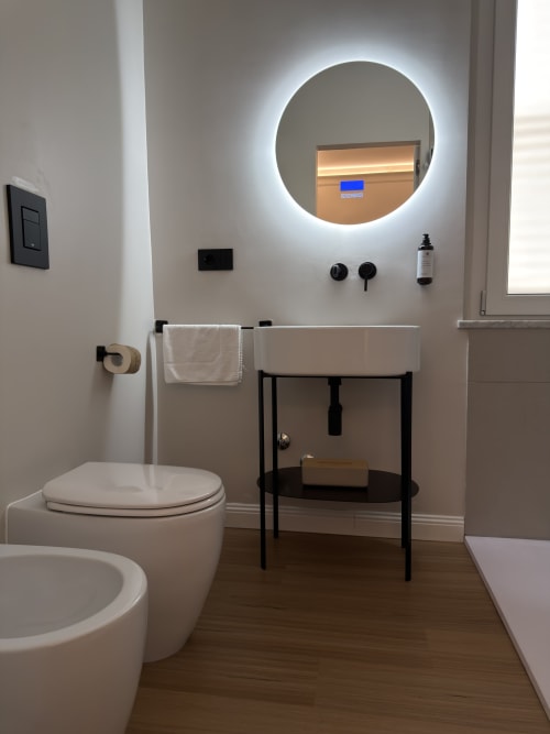 Bagno moderno di Maison Riviera con specchio rotondo illuminato, wc bianco, bidet, lavabo a colonna con struttura in metallo nero