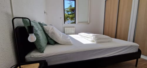 Chambre moderne avec lit en métal noir, literie blanche, coussins décoratifs verts et vue sur le jardin aux Terrasses des