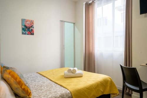 Quarto duplo iluminado na Madeirarooms Funchal com colcha amarela, janela com vista para a cidade e comodidades modernas