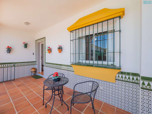 Terraza del loft Alhaurín con toldo amarillo, paredes de azulejos decorativos, macetas de flores, área de estar y ventana con rejas
