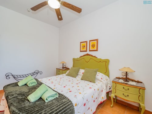 Dormitorio luminoso en villa Alhaurín Loft con cabecero amarillo, acentos verdes, ventilador de techo de madera y arte floral colorido.