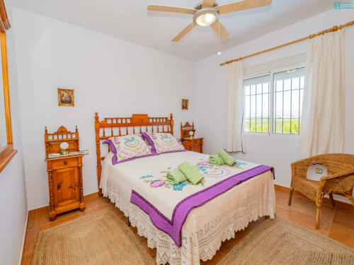 Dormitorio luminoso con cama de cuatro postes de madera, acentos púrpuras, ventilador de techo y amplias ventanas en la villa Alhaurín Loft