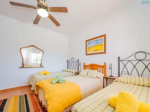 Dormitorio luminoso en Alhaurín Loft con tres camas, detalles amarillos, ventilador de techo de madera y alfombra de rayas coloridas