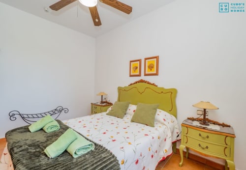 Dormitorio luminoso con cabecero amarillo ornamentado, ropa de cama floral, almohadas verdes, mesillas de madera y ventilador de techo