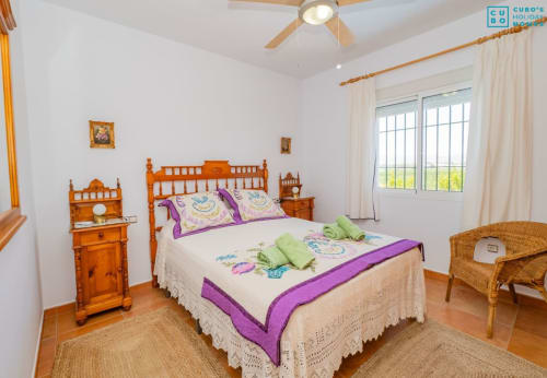 Dormitorio luminoso con cama de madera, acentos morados, sillas de mimbre y puertas de terraza en la villa Riad de Maruchi en Alhaurín de la