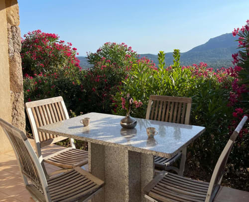 Terrasse de l'hôtel Vista D'oro à Monacia d'Aullène avec table en pierre, chaises en bois, fleurs roses en fleur et vue sur les montagnes