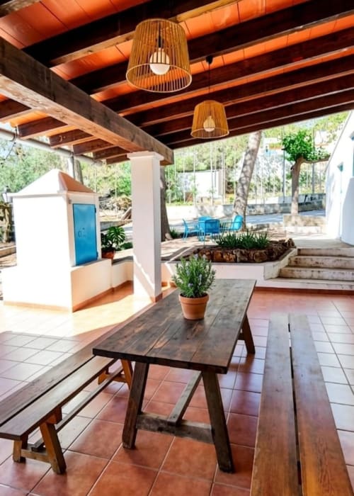 Patio cubierto de La Caseta de Ròtova con mesa de madera, bancos, plantas en macetas y vistas a la montaña