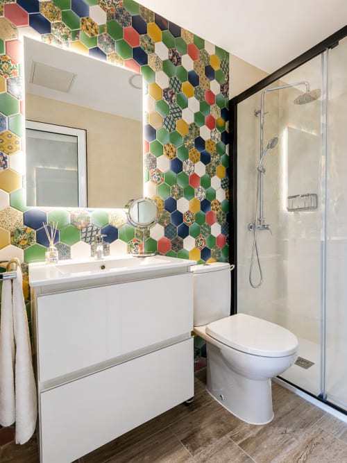 Baño moderno con pared de azulejos hexagonales coloridos, vanidad blanca, inodoro y ducha con mampara de cristal en Apartamento
