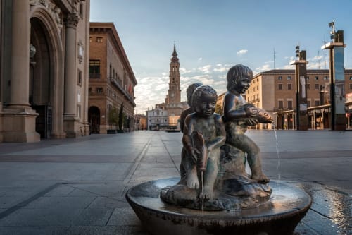 Estatua de bronce de niños en Plaza Square, Zaragoza, con arquitectura histórica y campanario de catedral al fondo