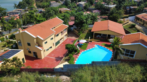 Vista aérea da pousada Equador Houses em São Tomé com piscina azul brilhante, pátio vermelho e edifícios amarelos circundados por