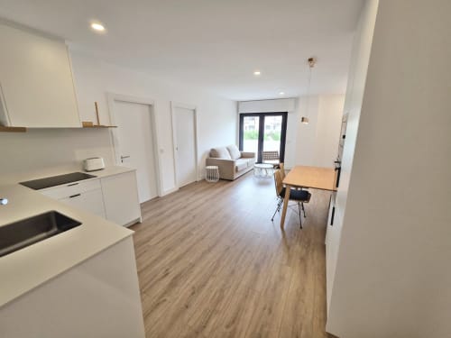 Apartamento moderno diáfano en BR Silgar Sanxenxo con cocina blanca, mesa de comedor de madera, sofá beige y balcón con puerta de cristal