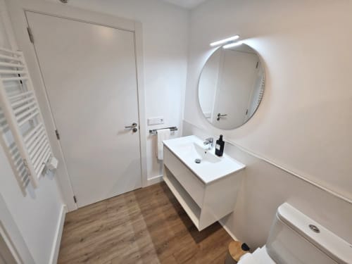 Baño moderno del apartamento BR Silgar en Sanxenxo con lavabo blanco, espejo redondo, inodoro y suelo de madera