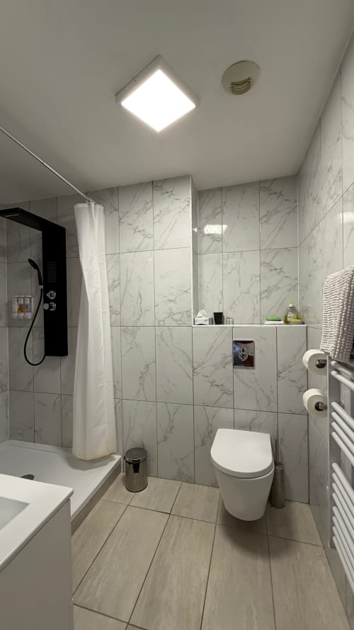 Salle de bains moderne de la Kimi Résidence Cannes avec carrelage marbre, douche à l'italienne, toilettes blanches, sèche-serviettes chauffant et plafonnier LED