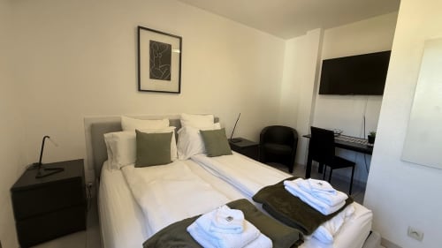 Chambre studio à la Kimi Résidence Cannes avec lit blanc, oreillers verts, TV écran plat, bureau et tableau paysager