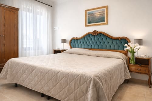 Camera elegante nell'hotel di Venezia con testata imbottita in velluto azzurro-verde, biancheria crema, comodini in legno e quadri con vedute marine