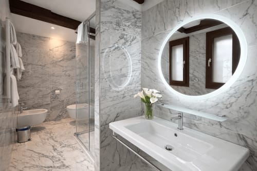 Bagno moderno in marmo con specchio rotondo illuminato, lavabo bianco e doccia in vetro all'Hotel Ariel Silva Venice