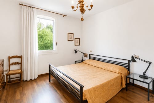 Camera luminosa a Palazzo Bonfiglioli con letto in metallo, biancheria dorata, pavimenti in legno e finestra con vista sul giardino a San