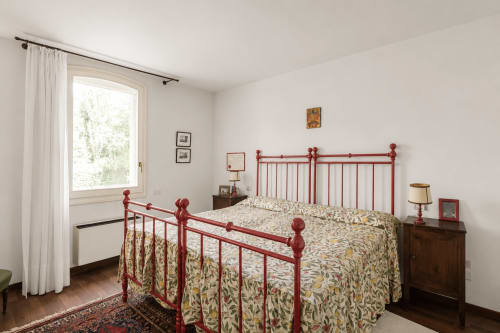 Camera luminosa a Palazzo Bonfiglioli con letto in ferro rosso, biancheria floreale, comodini in legno e ampia finestra