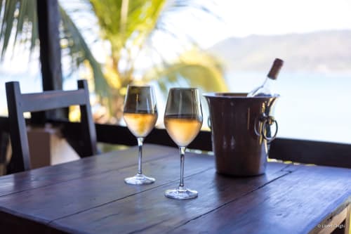 Deux verres de vin blanc sur une table en bois à la villa Madabay's Lodge de Nosy Bé avec vue sur l'océan et palmiers