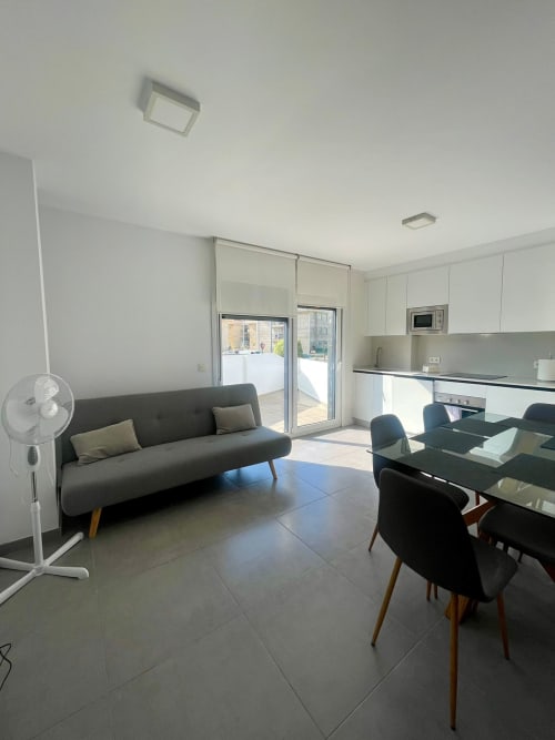 Moderna sala de estar con sofá gris, mesa de comedor, cocina y acceso a terraza en Baiona