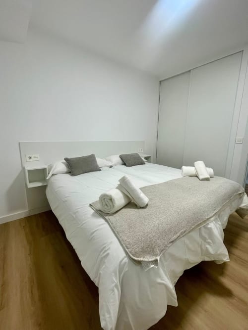 Dormitorio moderno en apartamento de Baiona con cama queen, ropa de cama blanca, almohadas grises, mesillas incorporadas y tragaluz