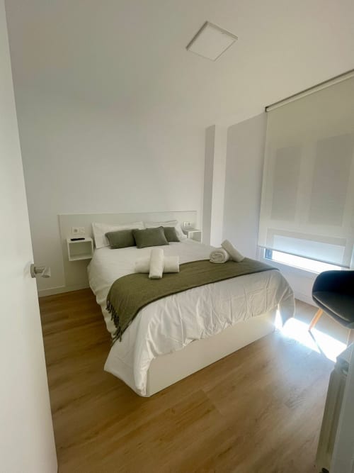Dormitorio moderno en apartamento de Baiona con cama queen, ropa de cama blanca, almohadas oliva, pisos de madera y paredes minimalistas blancas