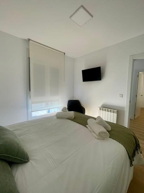 Dormitorio moderno en apartamento Bahía Rental de Baiona con ropa de cama blanca, TV montada en pared, radiador y acceso al baño