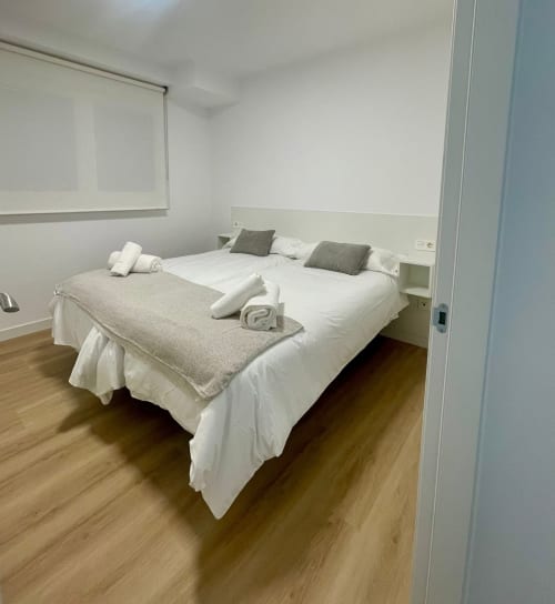 Dormitorio moderno en apartamento de alquiler en Baiona con cama doble, sábanas blancas, toallas enrolladas y decoración minimalista sobre piso de madera