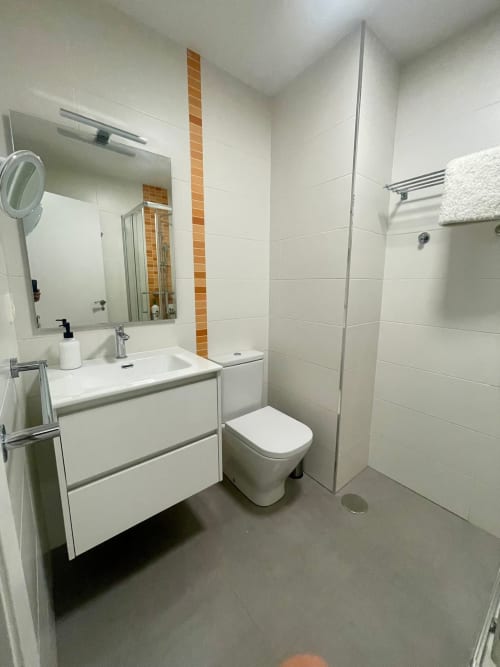 Baño moderno en apartamento de Baiona con vanidad blanca, inodoro, ducha a ras de suelo, azulejos naranjas y espejo
