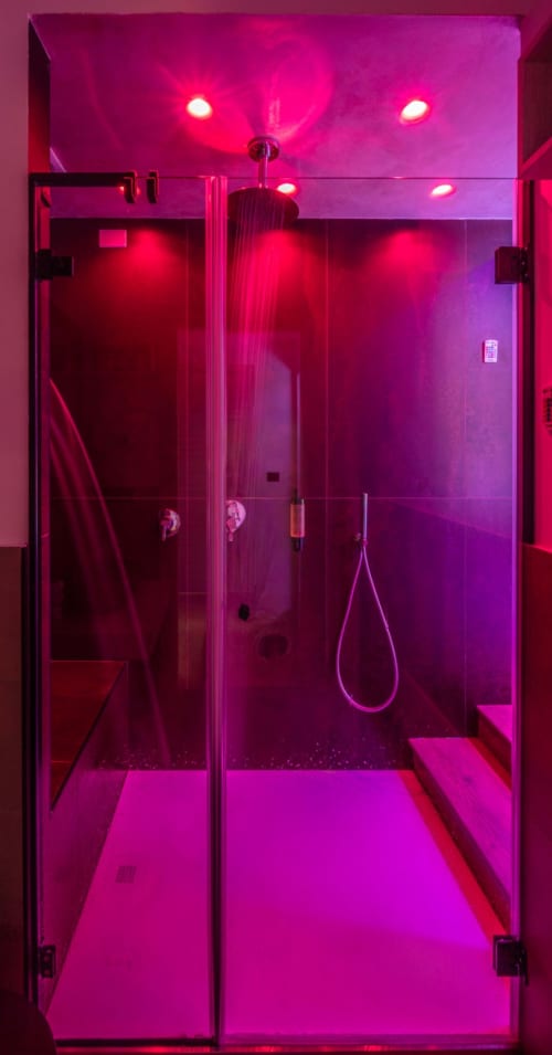 Modern bathroom with vibrant pink neon lighting, glass shower enclosure, rainfall showerhead, and marble floors in Suite